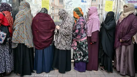 A line of women in traditional garb stand on the street outside a building