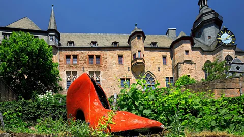 A large, red sculptural high-heeled shoe sits in a garden bed in front of a medieval sandstone castle (Credit: Alamy)