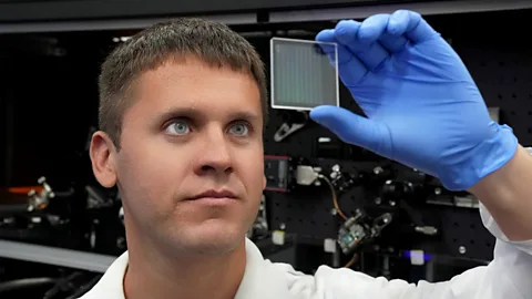Ilya Kazansky, CEO of Sphotonix wearing a white lab coat and blue latex gloves holds up a small square glass plate with grid markings on it to see through it (Credit: SPhotonix)