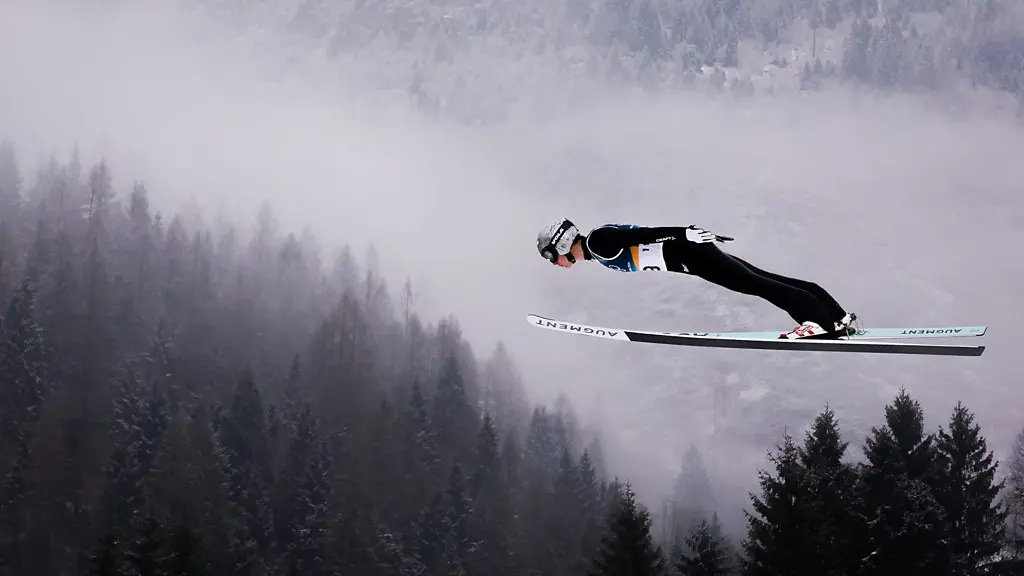 A ski jumper mid-jump upside-down, with his arms outstretched and snowy mountains in the background