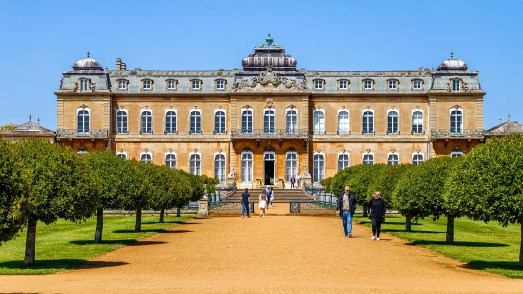 A man and woman together outdoors holding a film clapperboard that reads u2018Bridgertonu2019 along with scene and take numbers. The man is wearing period dress. There is a green metal fence or railings behind. It is sunny.