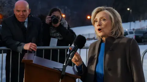  Hillary Clinton speaks to the press outside. She is wearing a blue top and a brown jacket