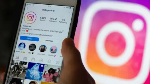 A smartphone screen showing social media platform instagram  and hand holding the phone with instagram logo in the background