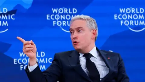 François-Philippe Champagne in navy suit and tie points his right hand towards the right with the blue backdrop that reads "World Economic Forum"