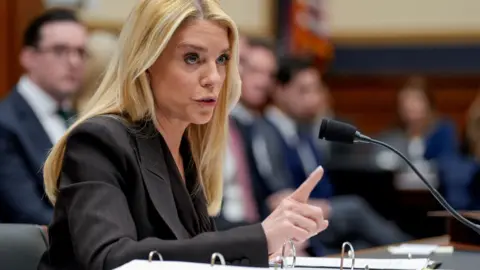 A woman in a dark suit with blonde hair sits in front of a mic pointing her index finger. 