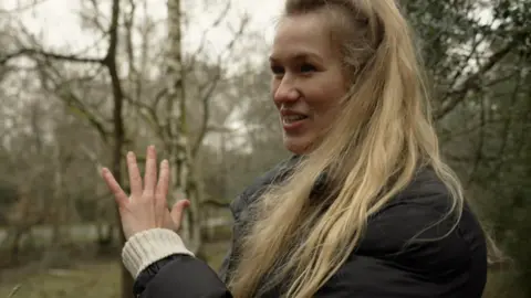 A woman with blonde hair in a dark jacket gestures in the forest