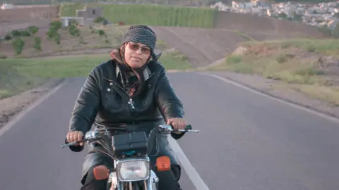 A woman rides a motorcycle along an empty road, with rolling hills seen in the background.
