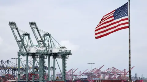 An American flag flies in front of shipping containers and cranes at the Port of Los Angeles on 26 September, 2025.