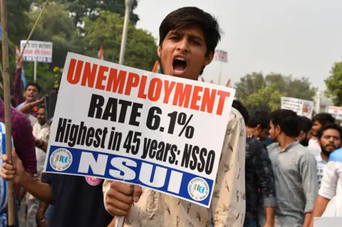 A young man wearing a white shirt holds a placard with slogans against unemployment and looks into the camera while shouting. Behind him are other protesters