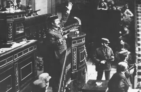 Coup leader Antonio Tejero Molina in police uniform stands at the top of stairs beside a lectern in the Spanish parliament brandishing a handgun in his right hand and with his left hand raised. Other armed men stand around him and in the distance MPs can be seen cowering down.