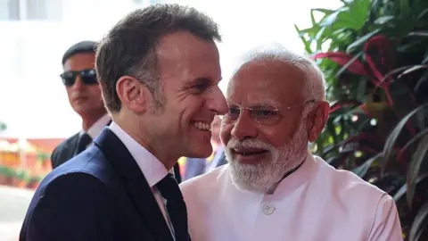 French President Emmanuel Macron (L) wearing a black blazer and tie over a white shirt, and India's Prime Minister Narendra Modi, wearing a white bandhgala jacket, sharing a warm moment as they greet each other upon the former's arrival in India's Mumbai city for a bilateral meeting on 17 February. Behind them there's another person wearing a black suit and sunglasses.