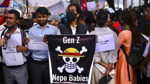 A man in a black facemask holds a poster saying Gen Z Nepo Babies, with a skull and crossbones in the middle. Around him other protesters stand, holding printed signs. Flags can be seen being waved in the background