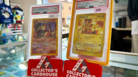 Two Pokemon cards stood upright on a counter in Collector's Cardhouse branded displays. One is a Dark Charizard card, the other is Pikachu card with Japanese text.
