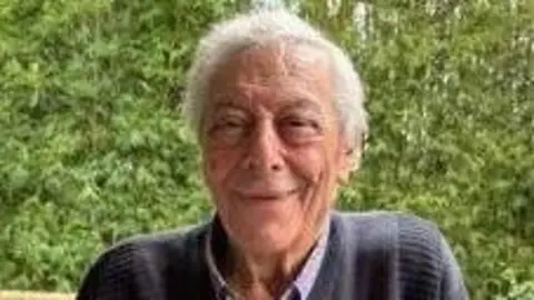 An elderly man with grey hair and wearing a dark blue jumper smiles at the camera