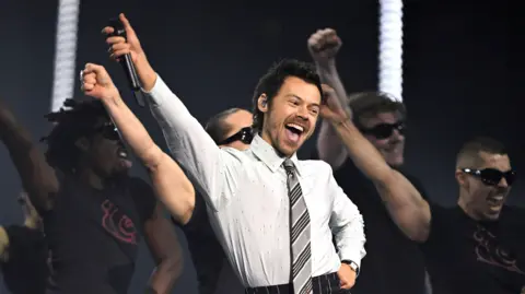 Harry Styles performs at the Brit Awards. He is wearing a grey office shirt and tie combination.
