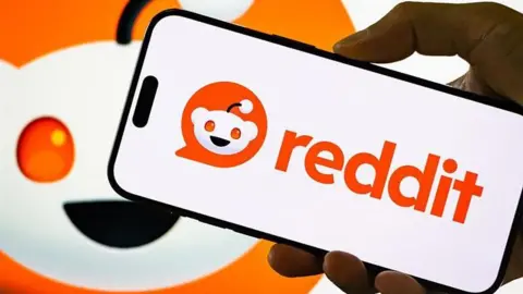 Reddit logo displayed on a smartphone held by hand, with a larger version of the company's logo behind it.