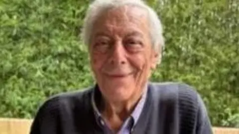 An elderly man with grey hair and wearing a dark blue jumper smiles at the camera
