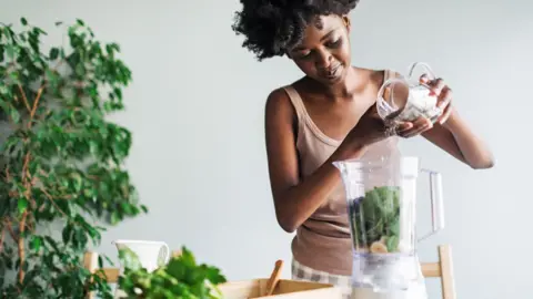 Stock photo shows a woman adding chia seends into a smoothie at home, there is a plant in the background.