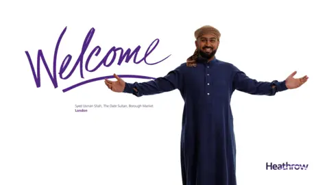 Syed Usman Shah features in a Heathrow welcome poster. He is wearing a traditional blue robe and brown turban and is smiling, holding his hands out wide. The poster has the word 'Welcome' in large writing. Beneath that it says: Syed Usman Shah, The Date Sultan, Borough Market, London