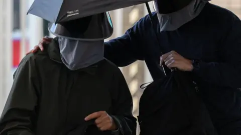 Two men dressed in black jackets and wearing tinted hats and face masks. They are sharing an umbrella held by one of them.