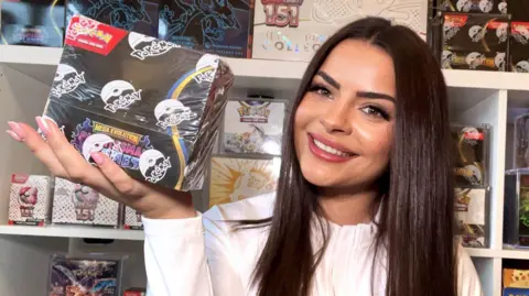 Monique has long dark brown hair and brown eyes. She is smiling holding a black box. Behind her are shelves of Pokémon merch, she;s wearing a white long sleeved jacket.