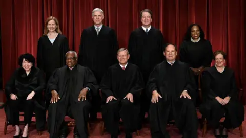 All nine justices pose for an official portrait in their black robes. Behind them is a dark red velvet curtain
