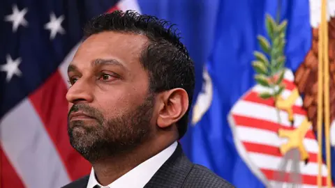 A close-up photo of Kash Patel in profile standing in front of a flag. He is wearing a suit and tie, and his expression is solemn.