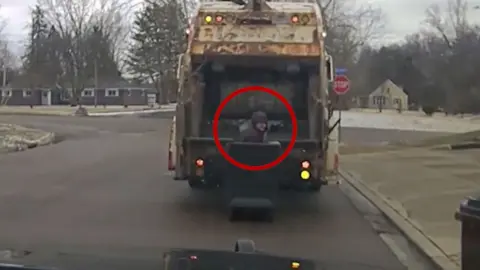 Man is hiding in a trash can with a garbage truck behind him.