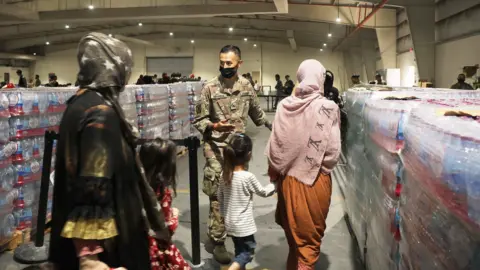 A soldier guides Afghan Special Immigrants. Pictured here are some women and children seen from behind,  into the dining facility, August 20, 2021 at Camp As Sayliyah, Qatar