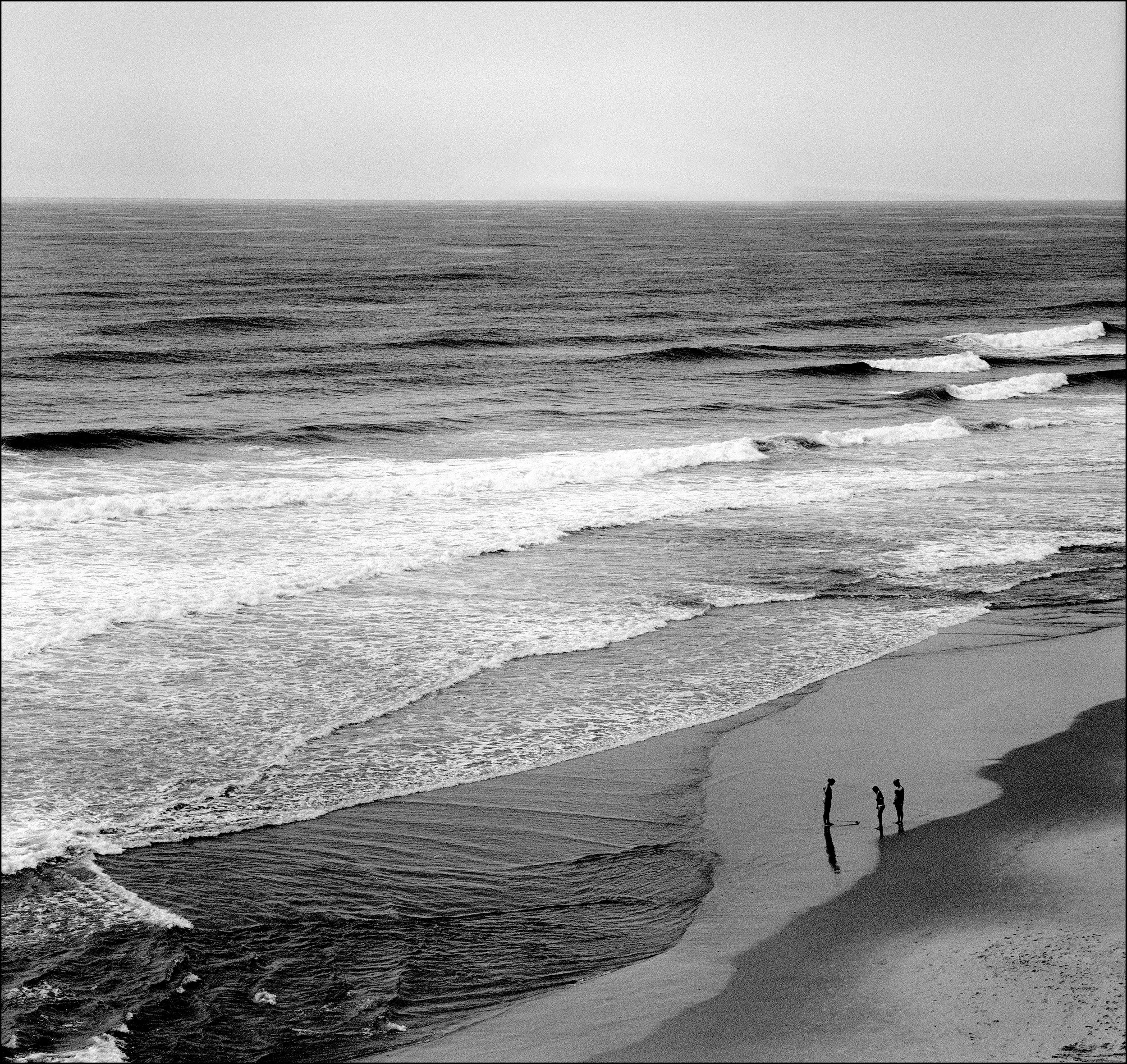 Black and white photograph of a beach with waves hitting the shoreline and three people standing on the sand.