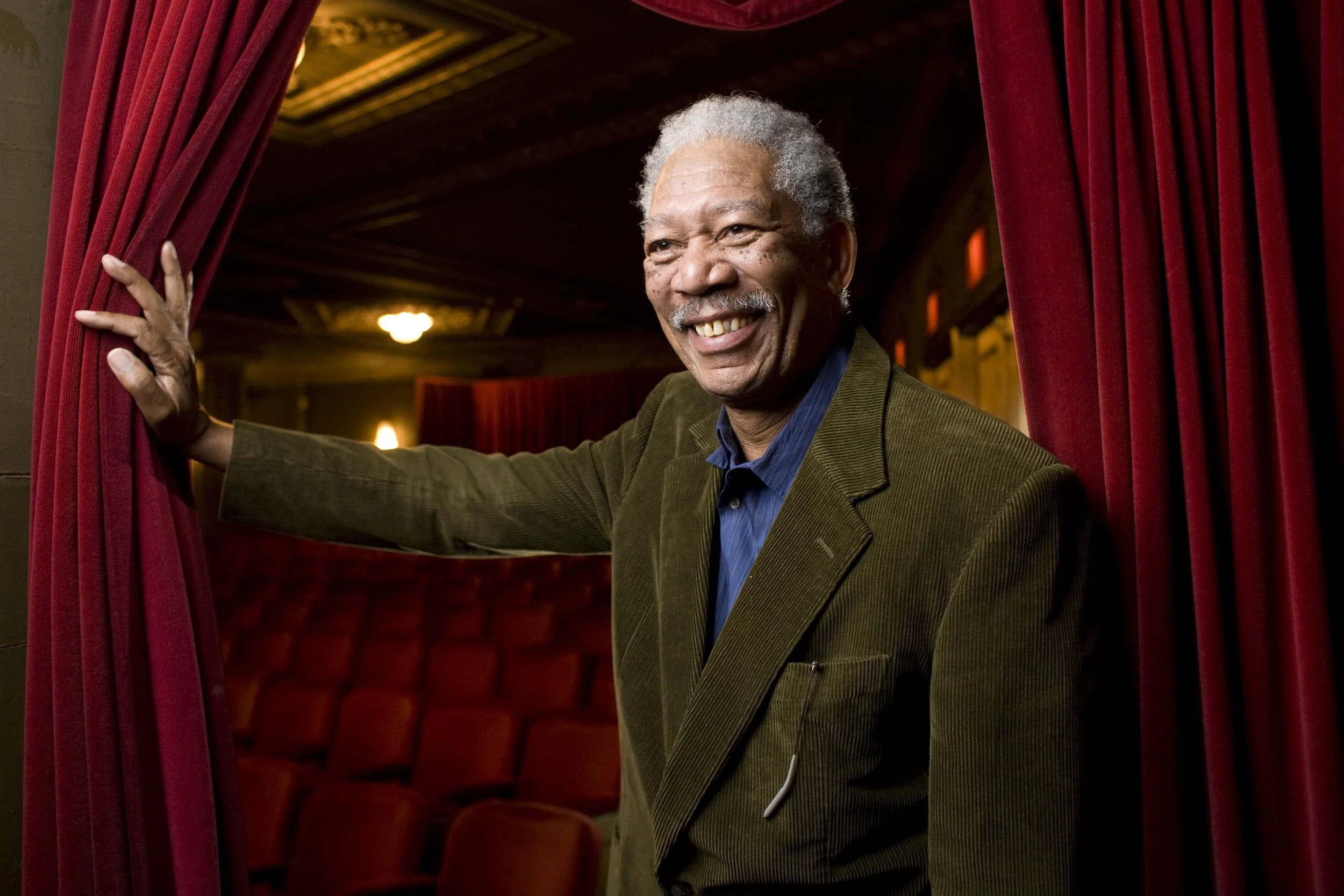 Smiling elderly man in a brown jacket holding back red theater curtains with an empty theater in the background.