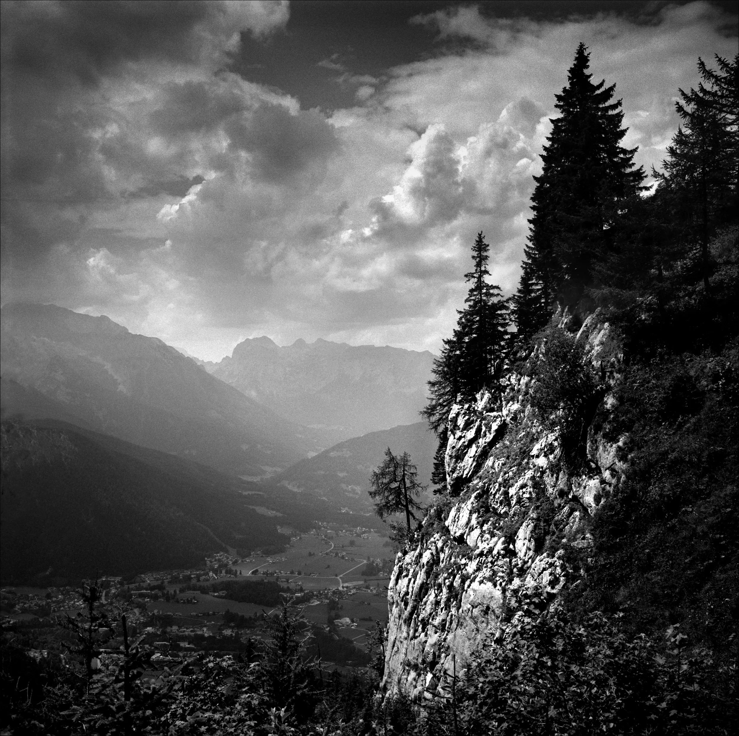 Black and white photo of a mountainous landscape with a rocky cliff on the right, tall pine trees on the cliff, distant mountains in the background, and a valley below.