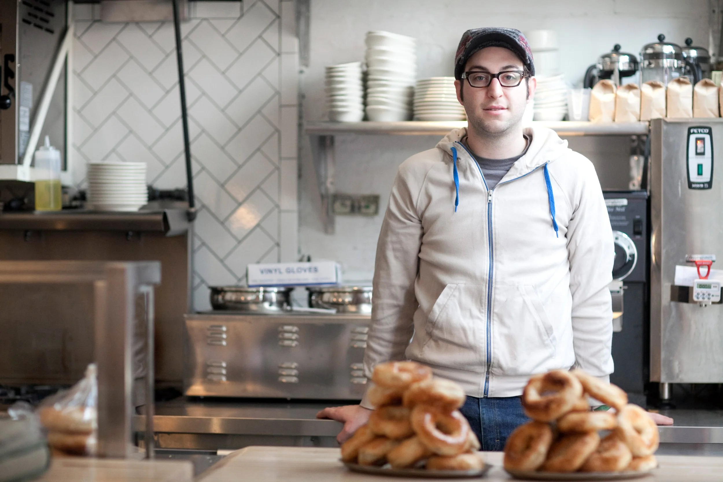 A young man in a hoodie and glasses standing behind a counter with plates and baked goods in front of him inside a cafe or bakery.