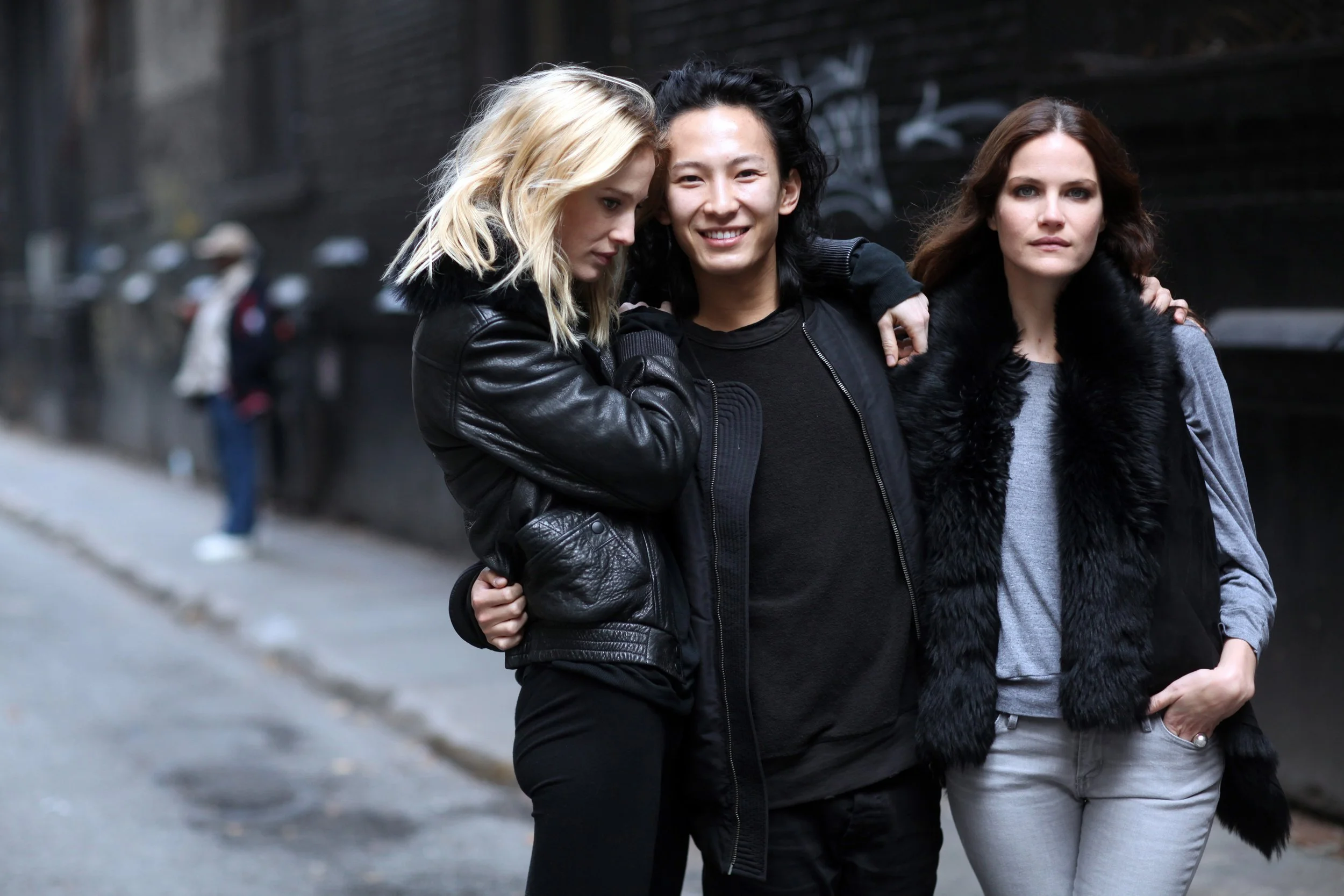 Three young women walk together on a city sidewalk, dressed in stylish black and gray clothing, with one smiling directly at the camera while the others look away or down.