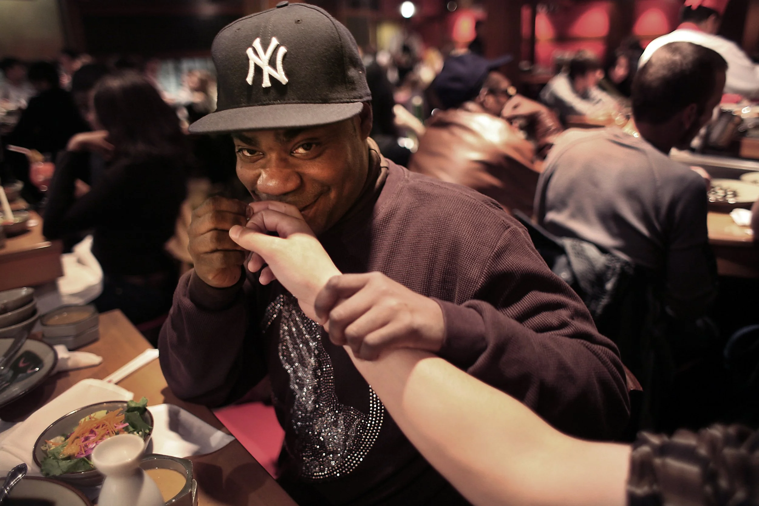 A man in a dark New York Yankees baseball cap holding hands with a person outside the frame, in a busy restaurant with other diners in the background.