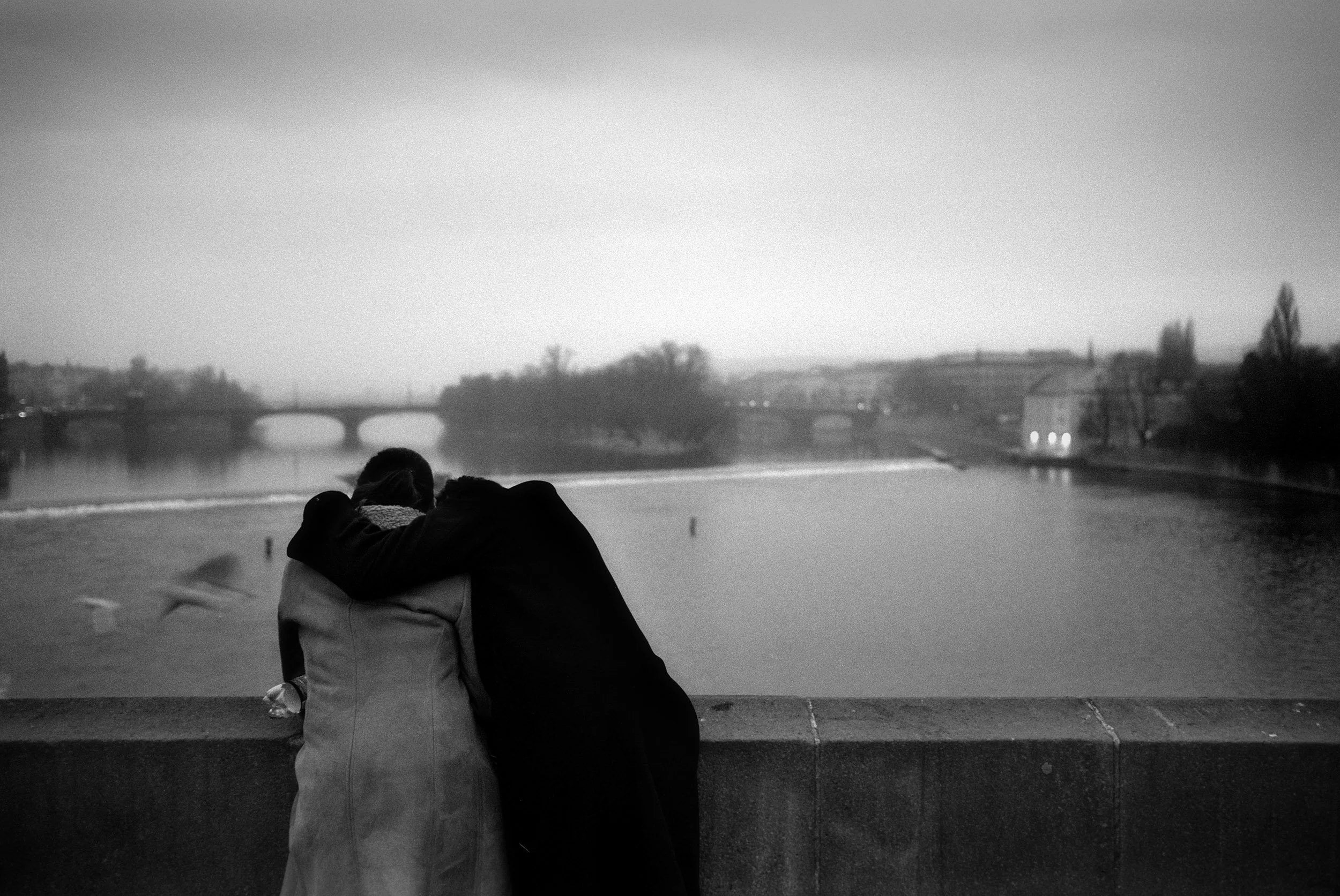 Two people with their arms around each other on a bridge, looking out over a river with boats and a cityscape in the background
