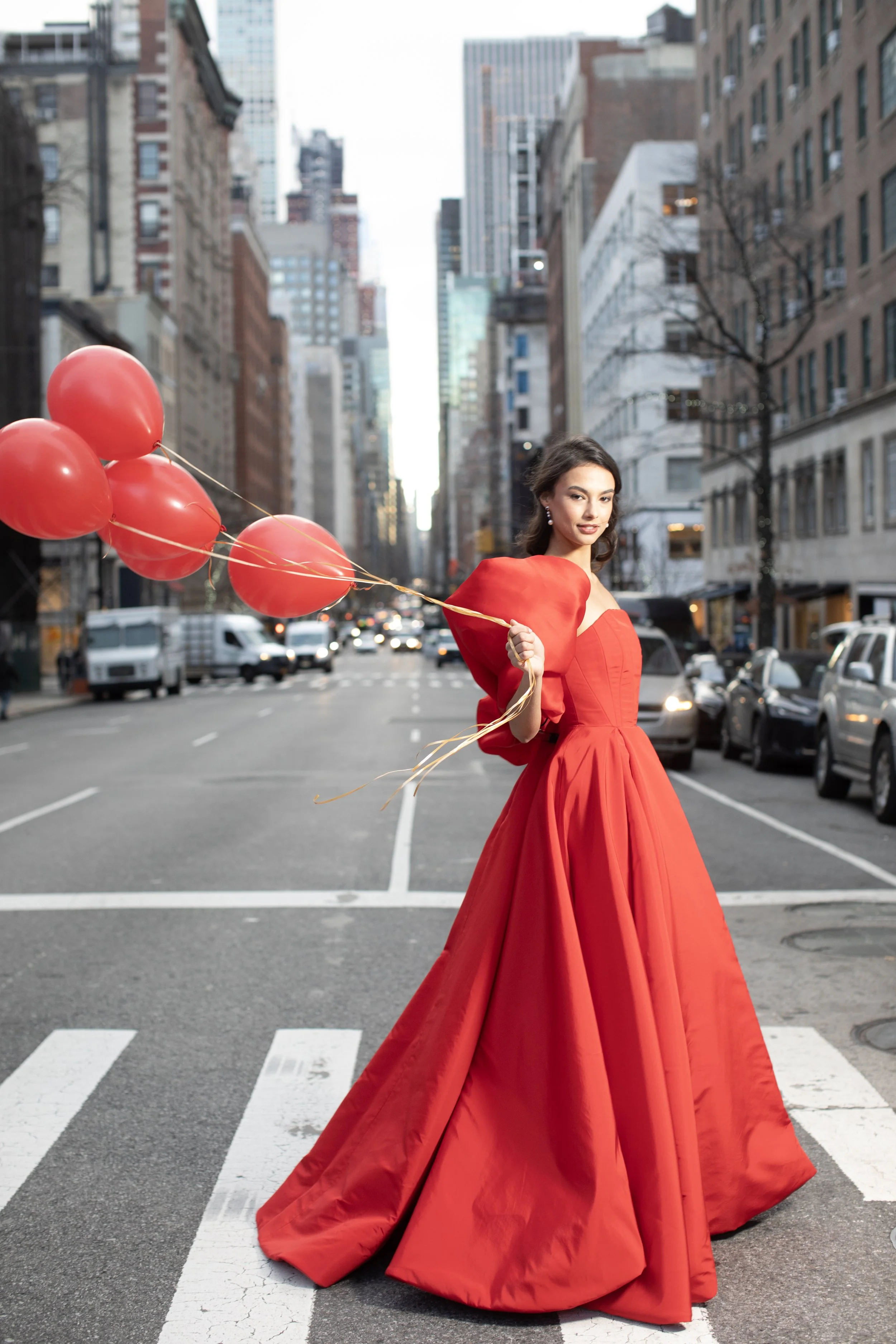 Woman in a red gown holding red balloons standing on a city crosswalk.