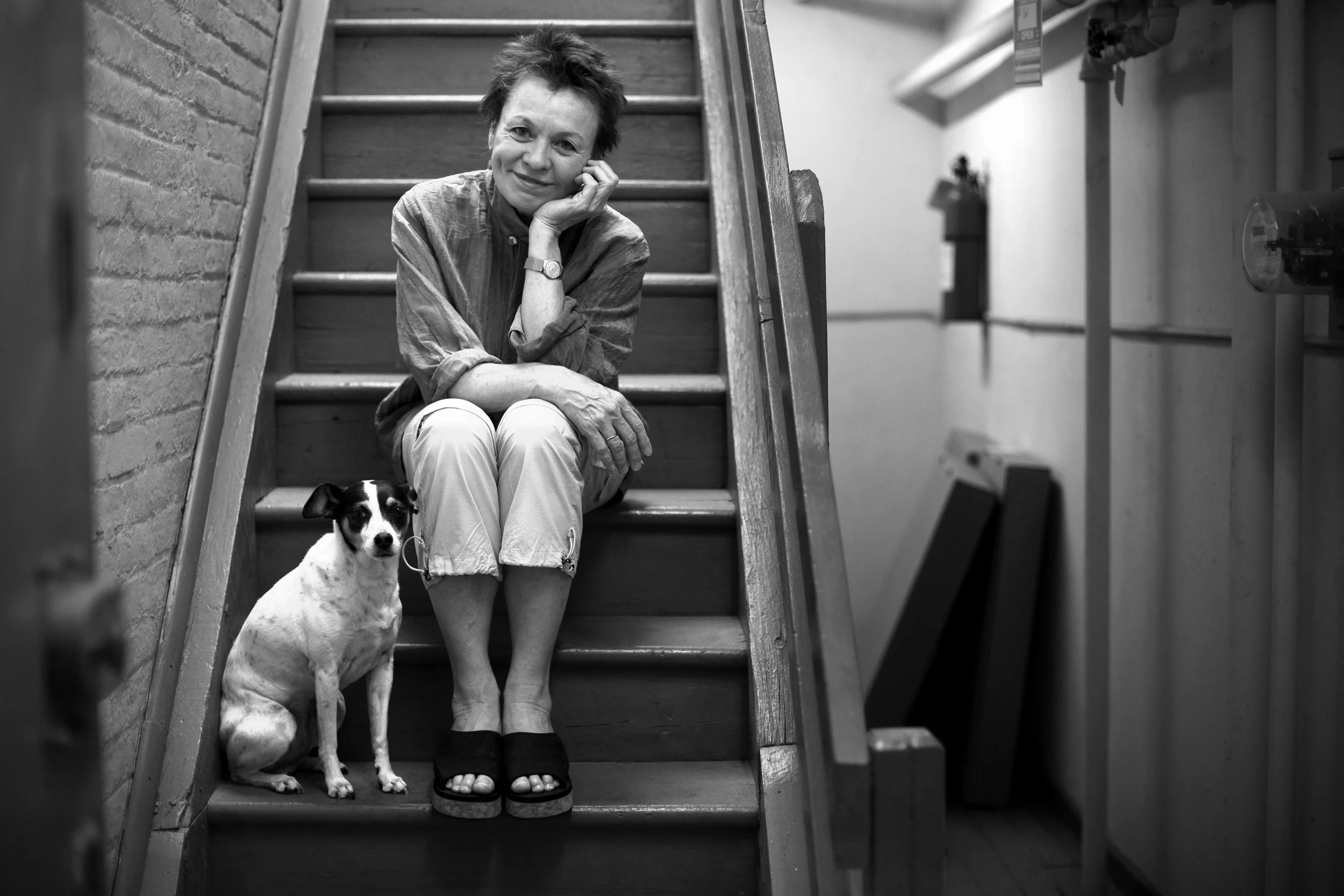 A woman sitting on wooden stairs with a small dog beside her. The woman is smiling and resting her head on her hand, wearing a casual shirt and pants. The scene is in black and white, with a brick wall on the left and a corridor on the right.