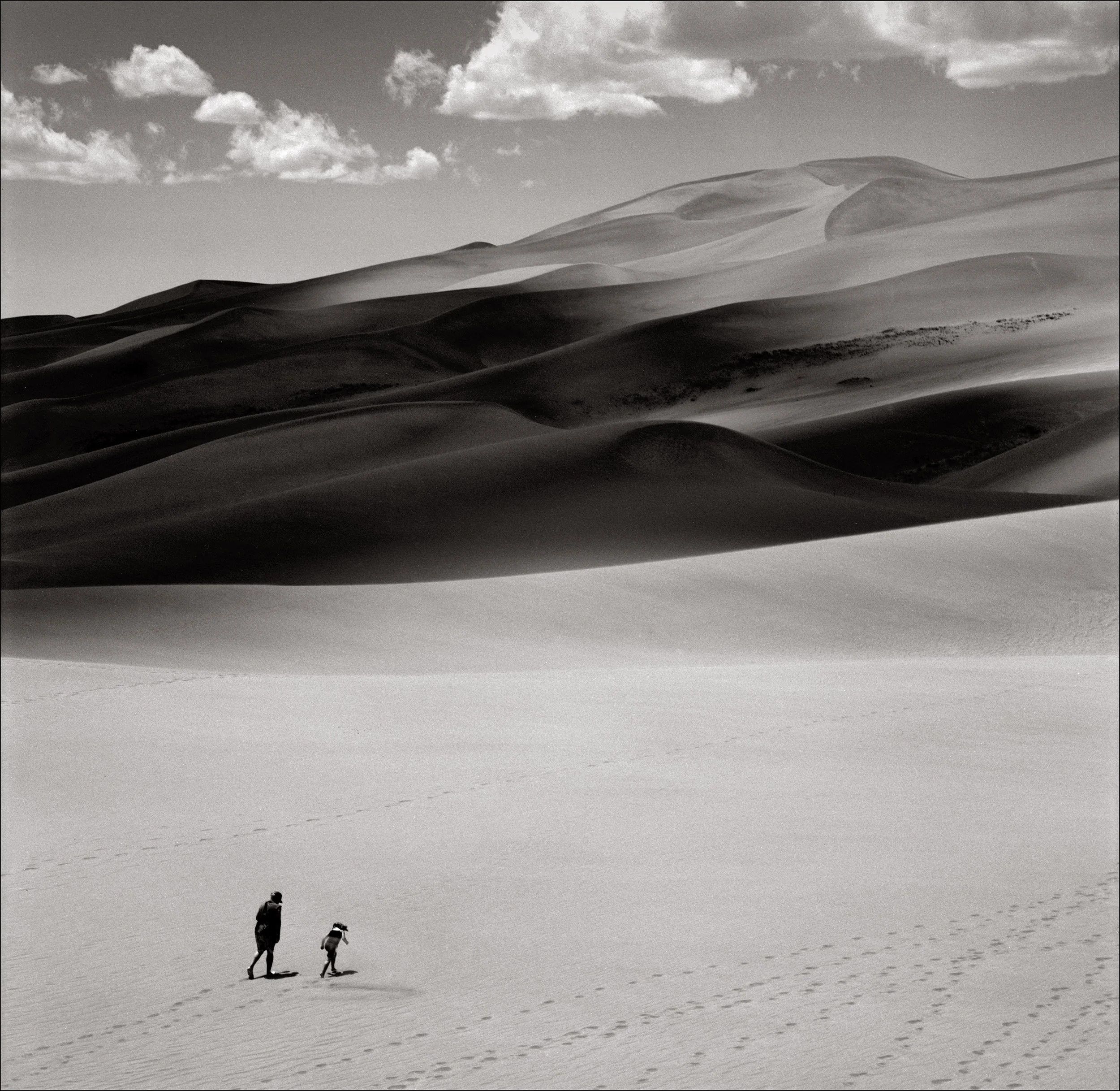 Two people, one adult and one child, walking across a vast desert with sand dunes and a cloudy sky in the background.