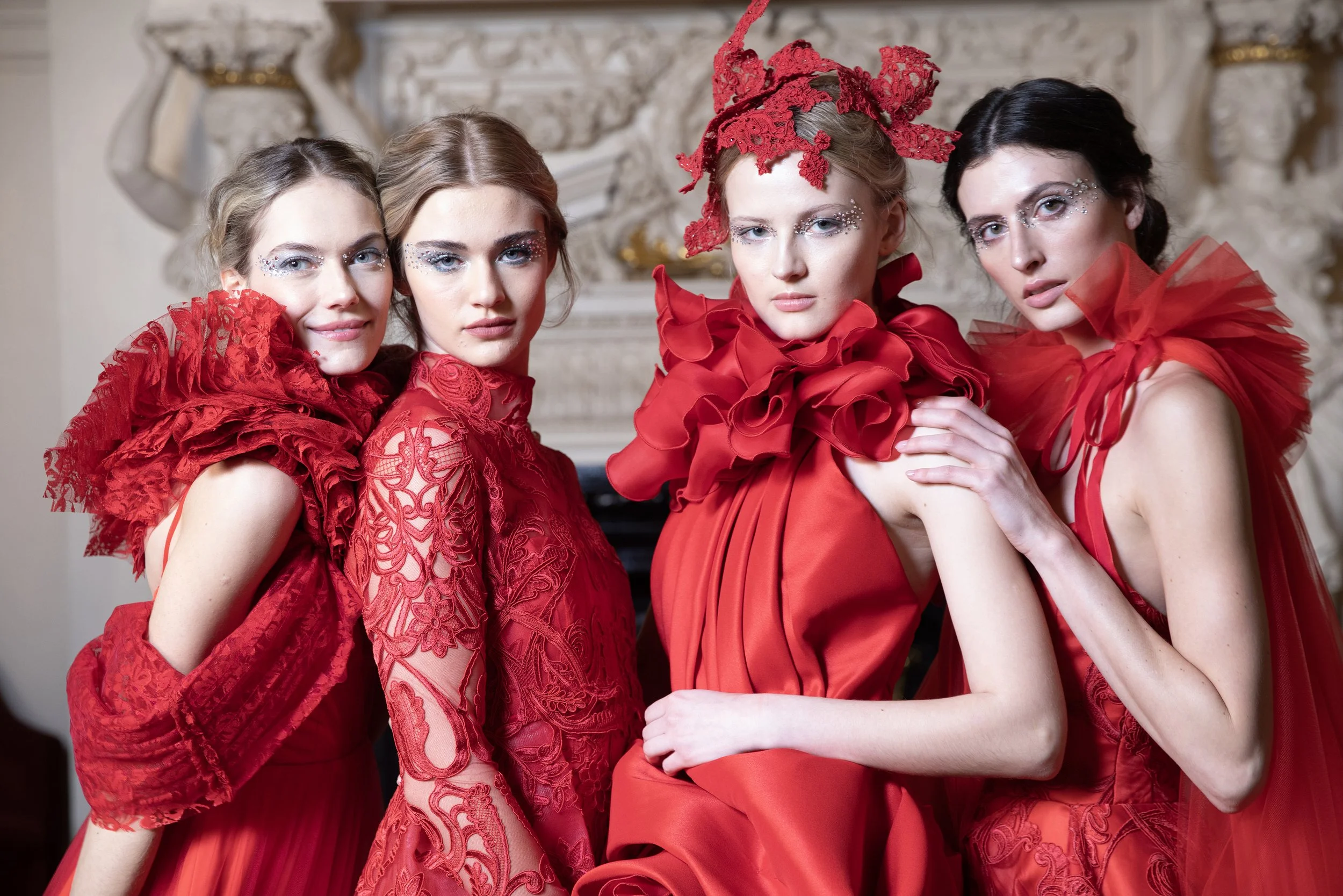 Four women dressed in elaborate red dresses with ruffled and lace details, standing close together in front of an ornate, classical interior background.