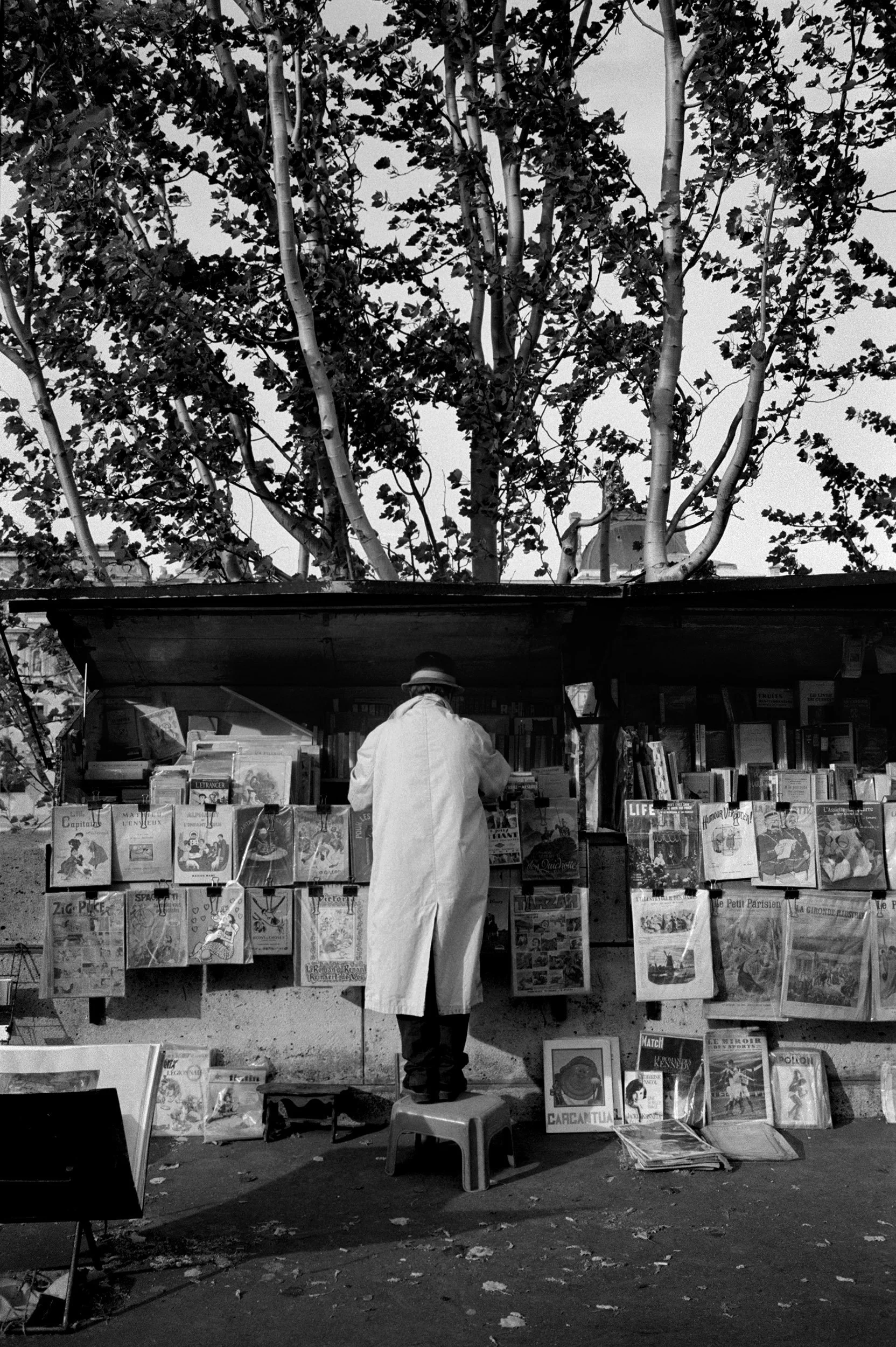 A person in a white coat and hat standing on a small stool, browsing paperbacks and magazines at an outdoor street book stall.