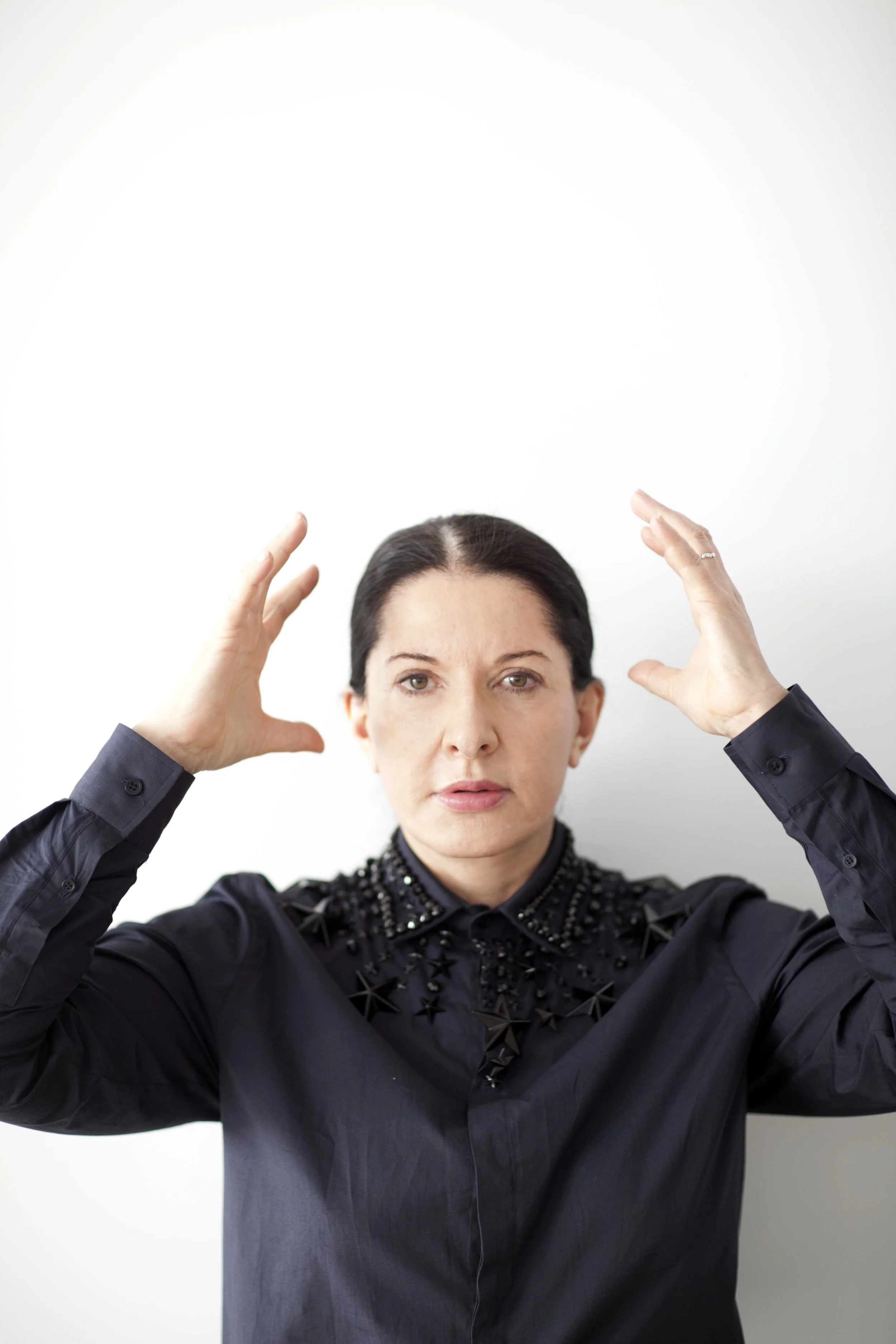 Woman with dark hair in a black shirt with star embellishments on the collar, raising her hands to the sides of her head, looking directly at the camera against a plain white background.