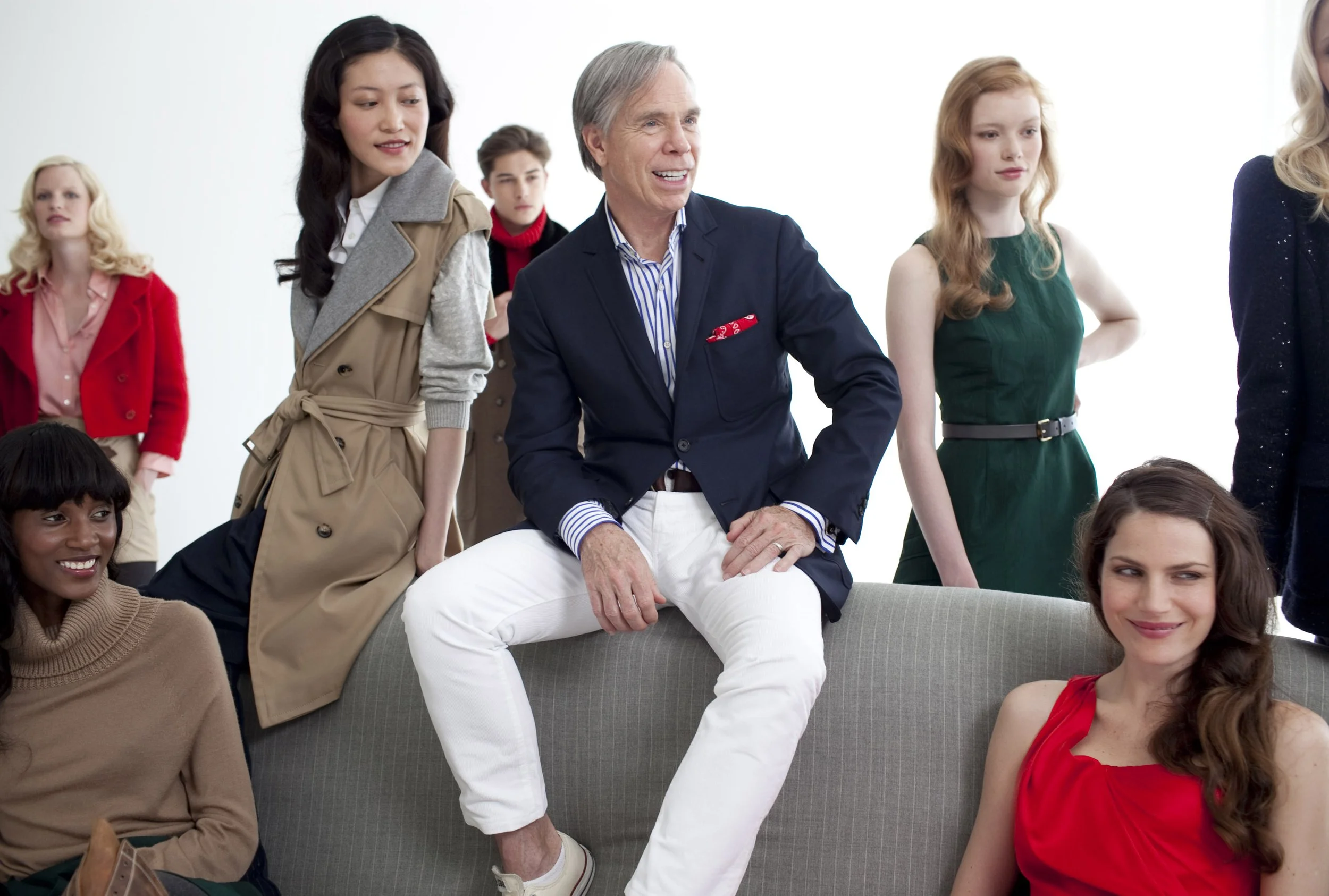 A diverse group of people at a social gathering, including women and men of various ages, with one man seated on a sofa in the foreground, smiling and engaging with others around him.