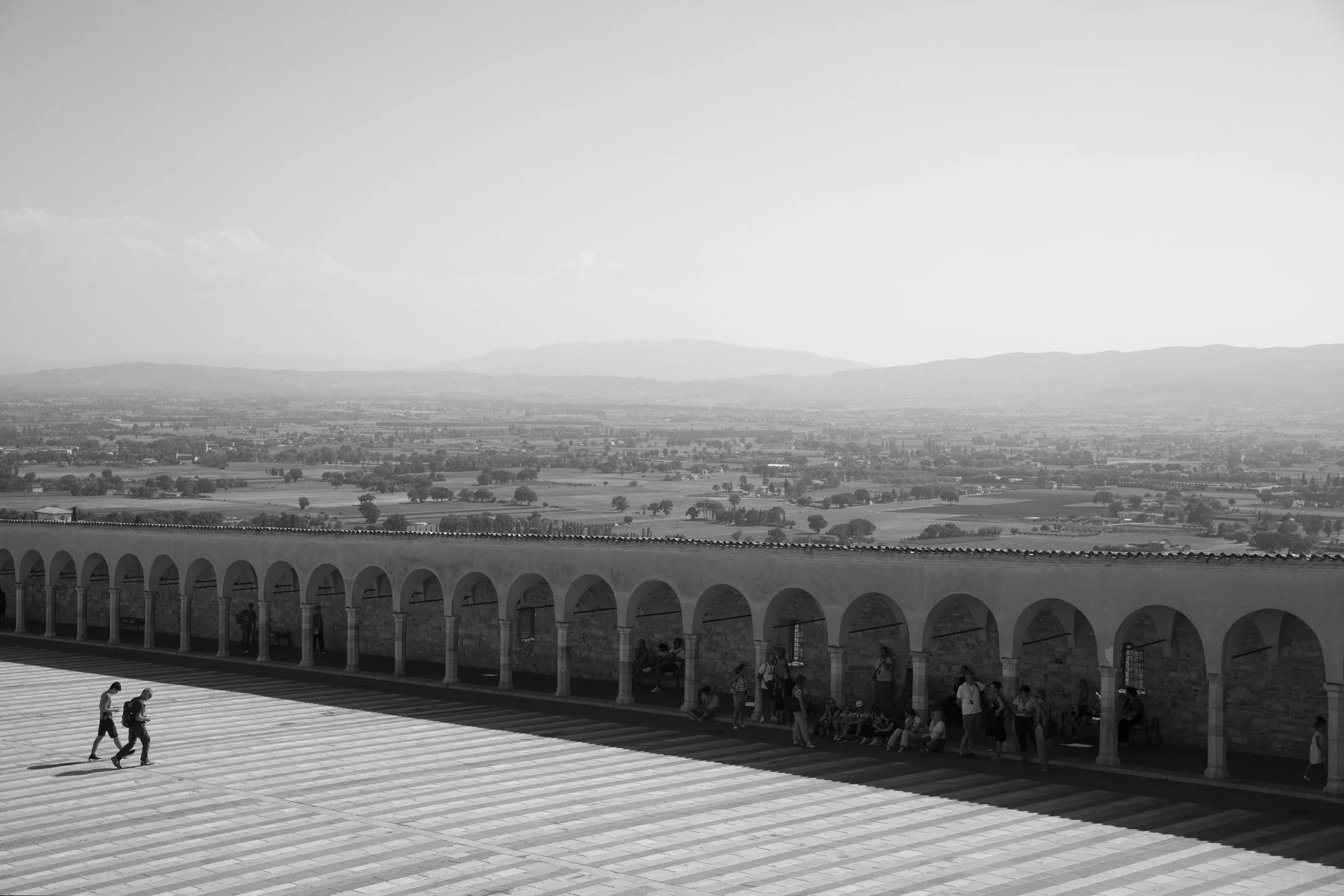 People walking and sitting under an arched walkway, with a vast landscape of fields and hills in the distance.