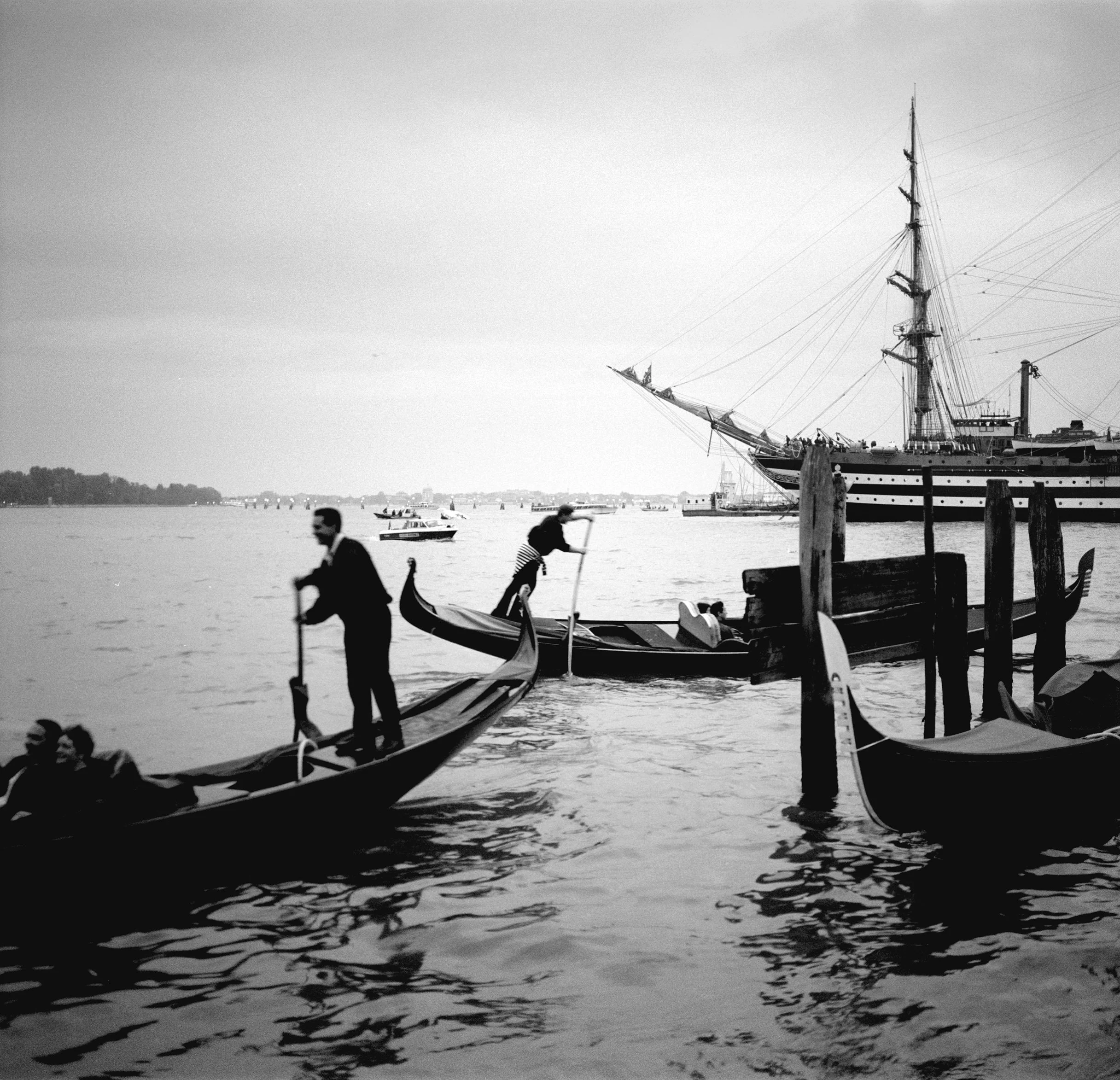 Black and white photo of gondolas and boats on a body of water, with a large ship in the background, and people riding in a gondola.