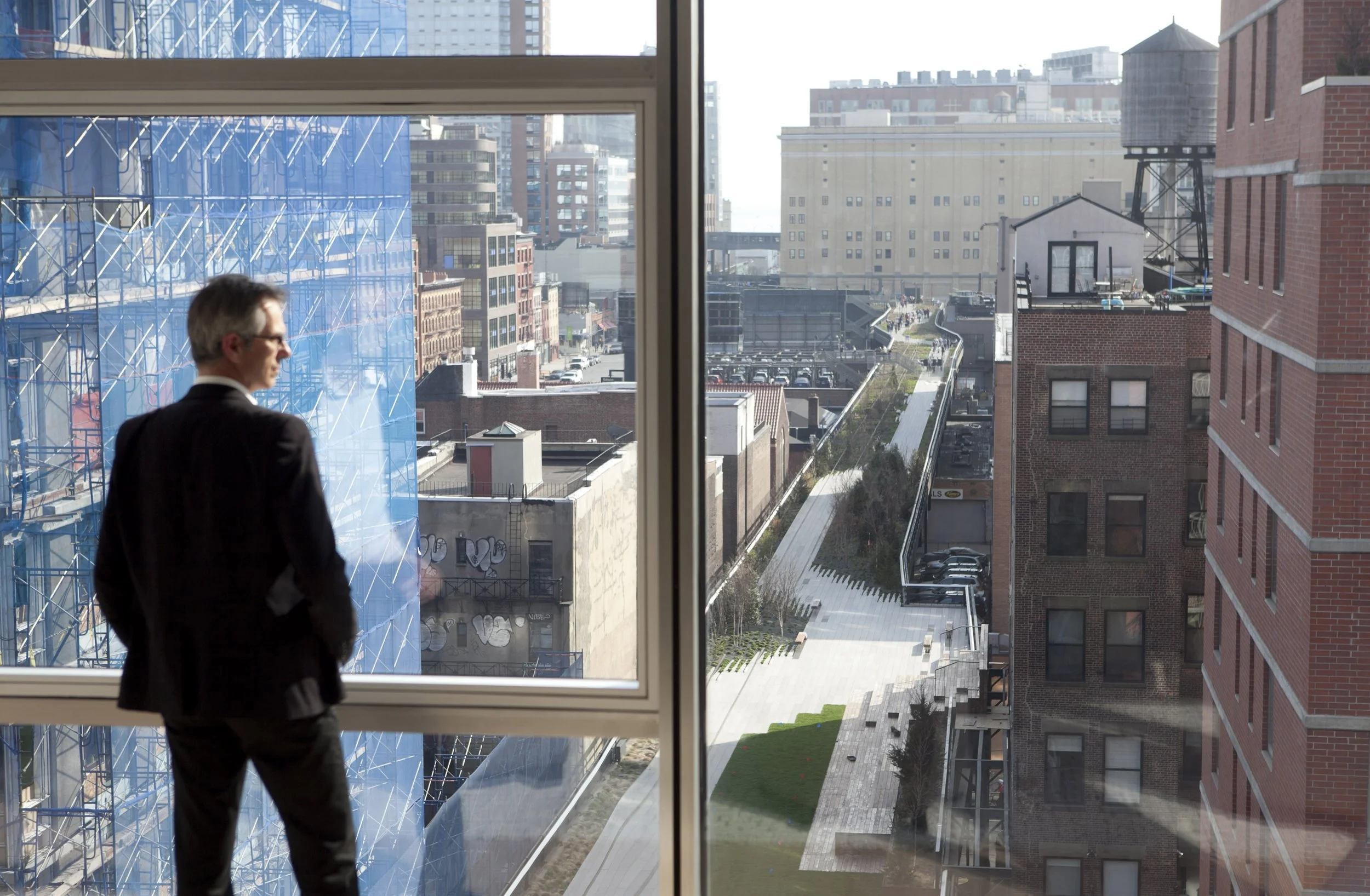 A man in a black suit looking out a window at a cityscape with modern and older buildings, some with graffiti, and a landscaped walkway outside.