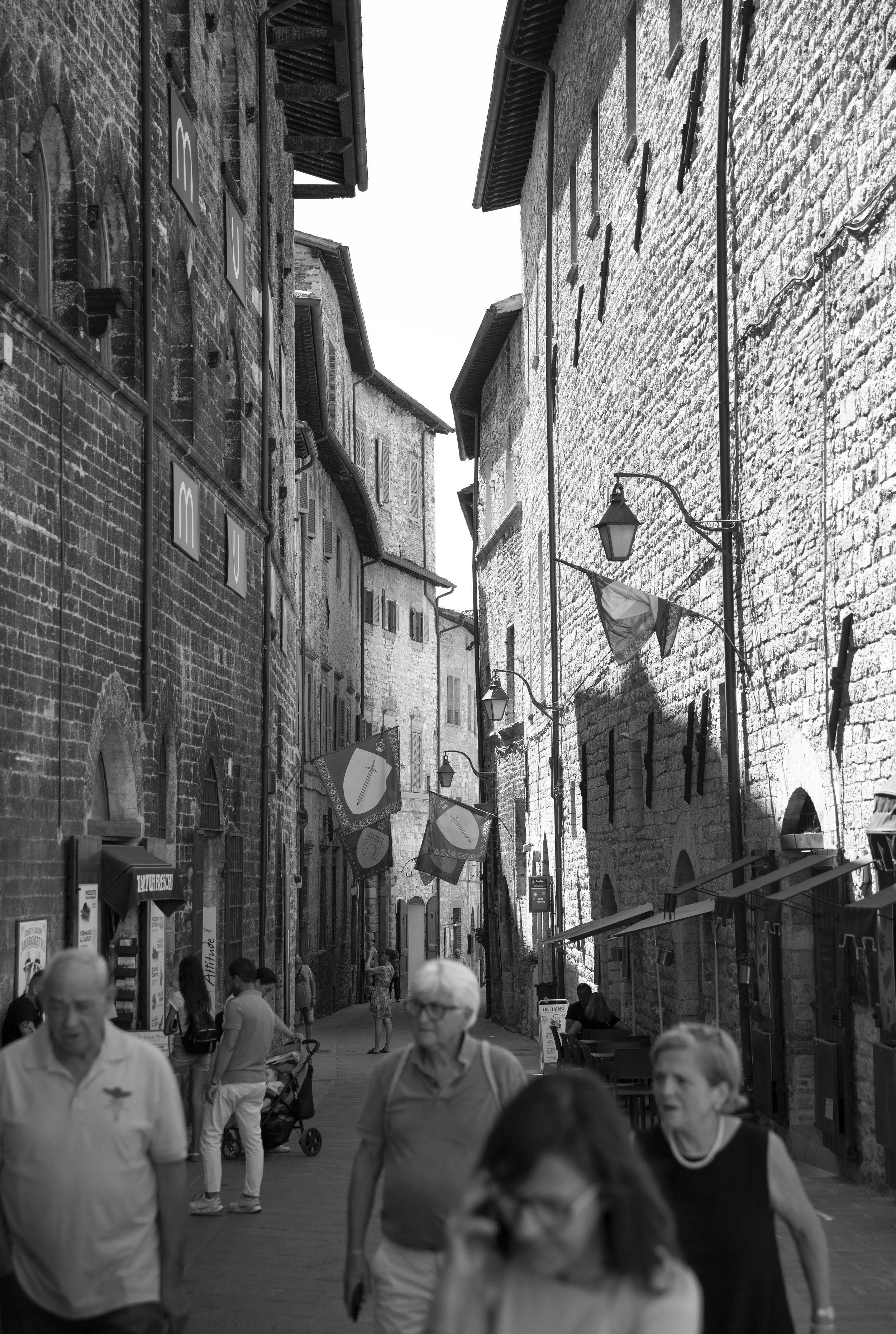 A narrow street with historic stone buildings on both sides, lined with hanging lanterns and flags. Several people are walking along the street, some in the foreground and others in the background.
