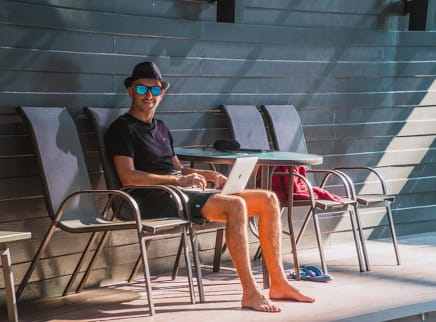 A man in shorts and a sun hat sitting with his laptop